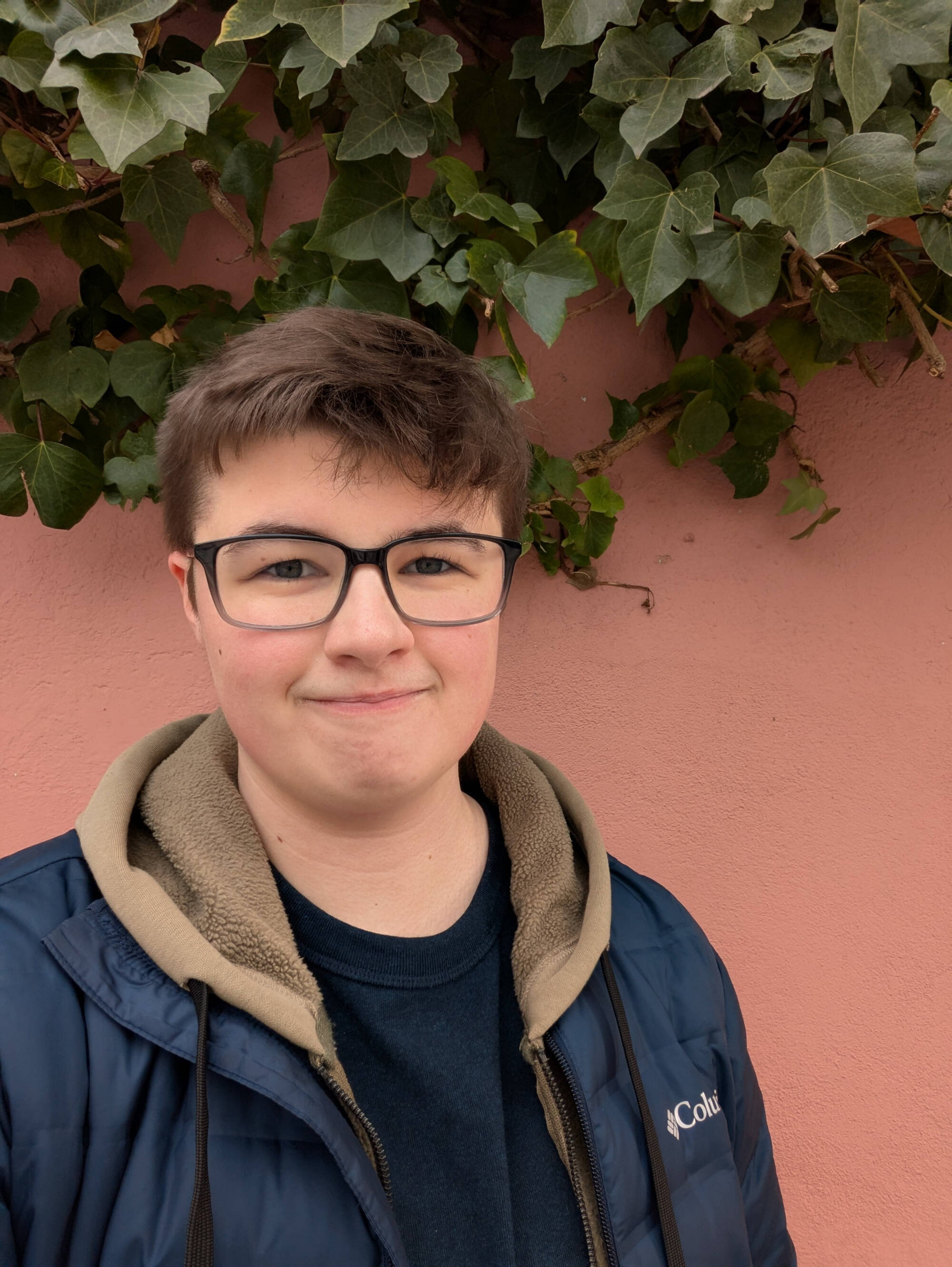 Alex Mulder wearing glasses and a blue coat in front of a pink wall with ivy leaves growing on it
